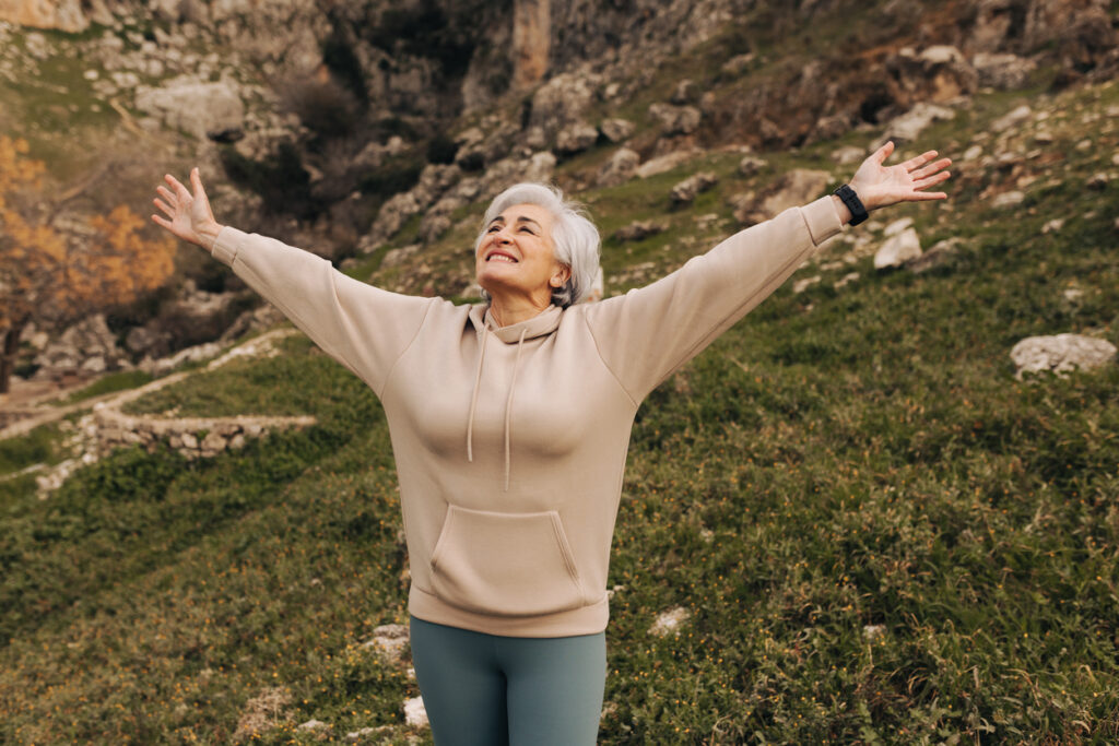 Happy senior woman celebrating with her arms outstretched while standing on a hill. Cheerful senior woman enjoying a leisurely hike outdoors. Woman enjoying recreational activities after retirement.