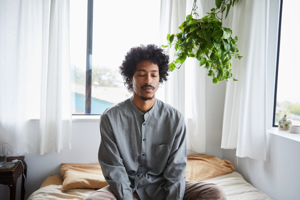 Young African American man sitting with his legs crossed and eyes closed on his bed during a meditation session