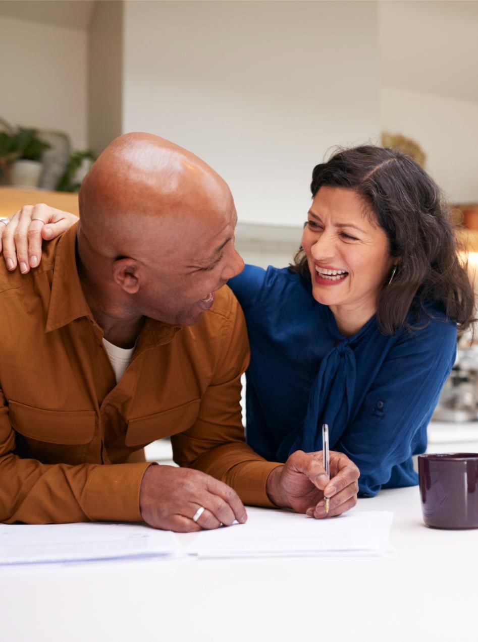 Mature Couple Reviewing And Signing Domestic Finances And Investment Paperwork In Kitchen At Home