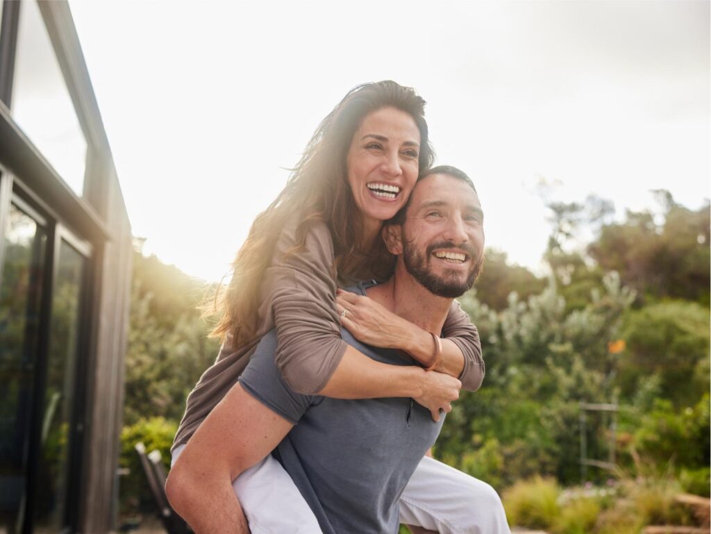 Man carrying his laughing wife on his back and laughing together while enjoying a sunny afternoon in their back yard