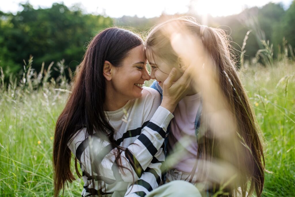 Beautiful mother with daughter, touching with foreheads and sitting in the grass at meadow. Concept of Mother's Day and maternal love.