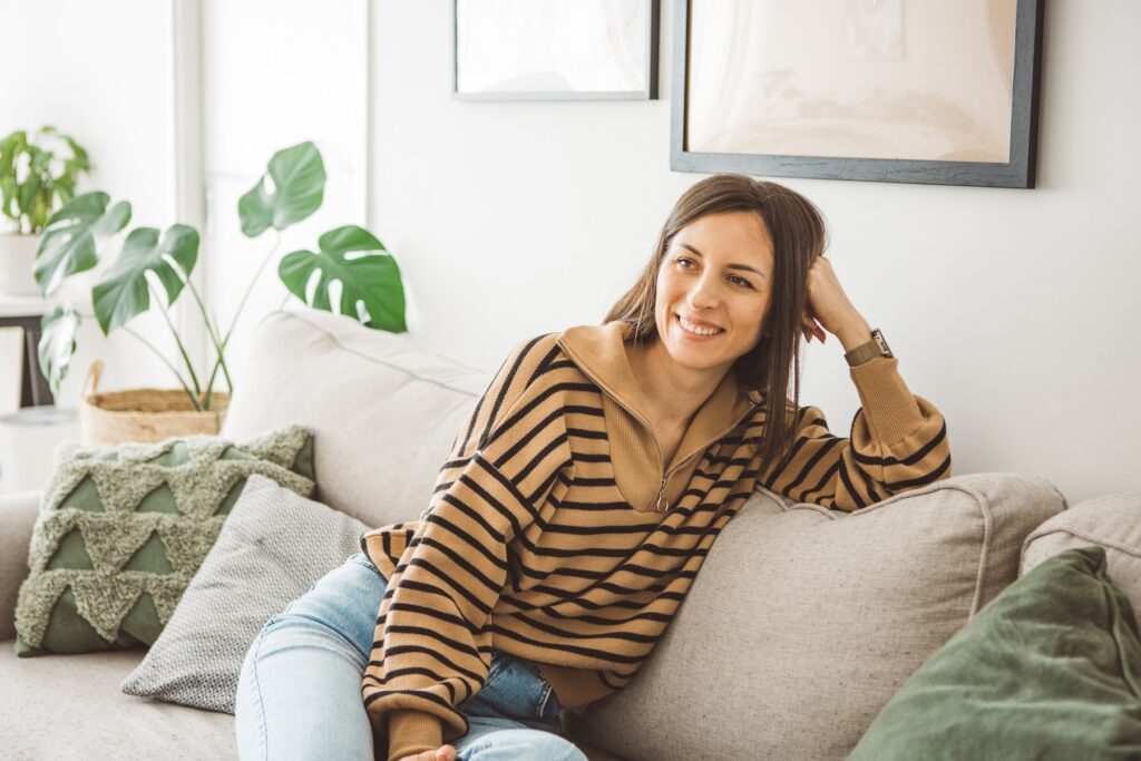 Smiling young woman using her mobile phone while drinking coffee sitting on sofa at home.