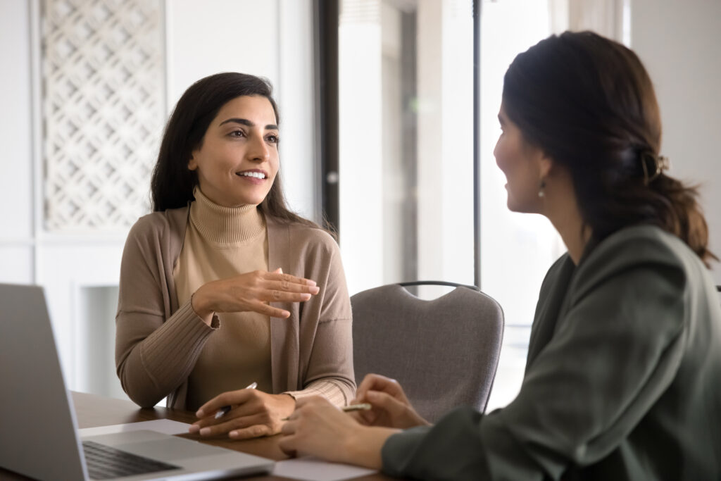 Positive attractive Hispanic business woman discussing project with colleague, talking at laptop. Two female coworkers brainstorming on tasks, chatting on work break. Mentor training intern
